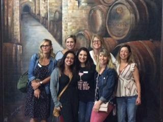 A group of 7 women smiling and standing in front of a mural of wine barrels